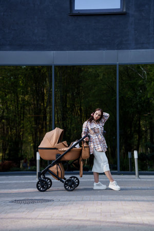 Portrait of a beautiful girl. Close-up photo of a beautiful girl in a plaid shirt and a cap. The girl stands near the brown baby carriage. Mother walking with a small childの写真素材