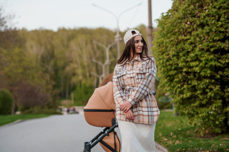 Portrait of a beautiful girl. Close-up photo of a beautiful girl in a plaid shirt and a cap. The girl stands near the brown baby carriage. Mother walking with a small childの写真素材