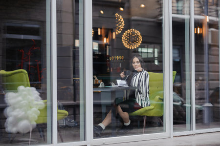 A beautiful young woman in a black dress sits in a cafe by the window and drinks coffee. Coffee...
