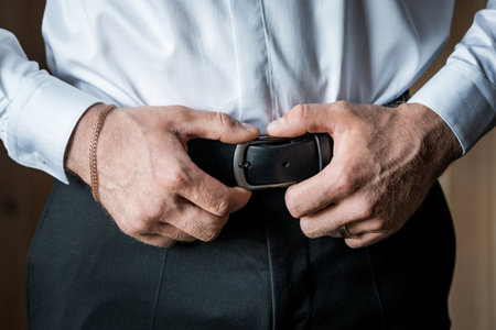 Close-Up of a Man's Hands Adjusting a Black Leather Belt Over Formal Attire.
