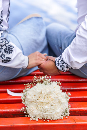 Romantic couple holding hands on a vibrant red bench with a beautiful bouquet of white flowers in the foreground.の写真素材