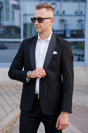Elegant Man in Tailored Black Suit with Sunglasses and Stylish Watches Posing Outdoors.
