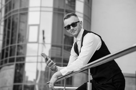 Stylish Businessman in Sunglasses Posing with a Smartphone Against a Modern Urban Background.の写真素材