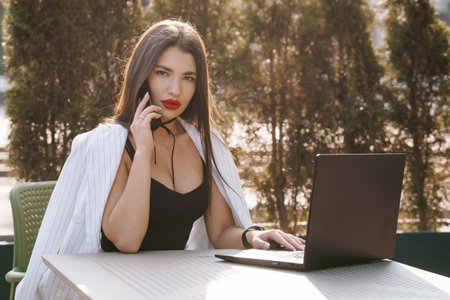A Confident Businesswoman Engaging on a Phone Call While Working on a Laptop Outdoors.の写真素材