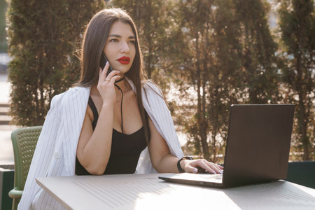 A Stylish Businesswoman Engaged in a Phone Call While Working on Her Laptop Outdoors.の写真素材