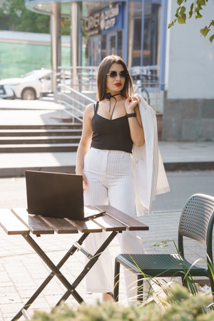 Young businesswoman in stylish attire standing beside a laptop on an outdoor table in a modern urban setting.の写真素材