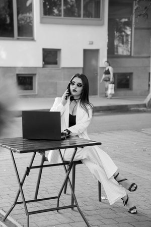 A Stylish Businesswoman Engaged in a Phone Call While Working on a Laptop Outdoors.の写真素材