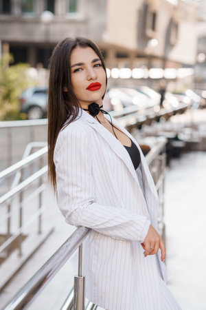 Stylish woman confidently posing outdoors in a chic striped suit with a bold red lip and a flower choker accessory.の写真素材