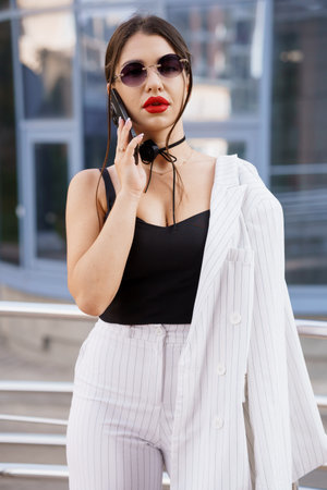 Stylish Businesswoman in Sunglasses and Striped Suit Holding Phone Outdoors.の写真素材