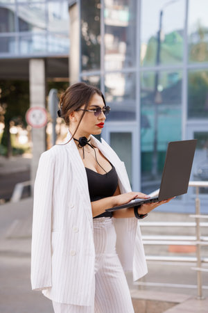 Stylish businesswoman in urban setting using laptop with sunglasses and sleek attire.の写真素材