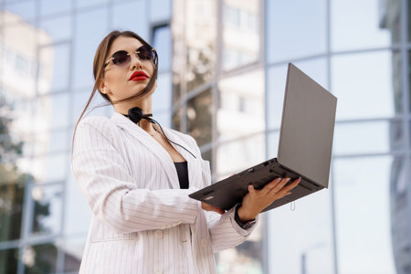 Confident Businesswoman in Striped Suit Holding Laptop Against Modern Glass Building.の写真素材