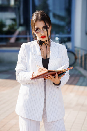 Stylish businesswoman jotting down notes in a notebook outdoors with confidence and elegance.の写真素材