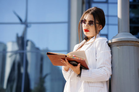 A Stylish Young Woman in Striped Blazer Reading a Notebook Outside a Modern Glass Building.の写真素材