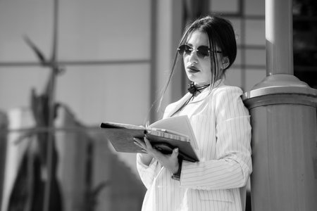 Stylish Young Woman in Pinstripes with Sunglasses and Notebook by a Modern Building.の写真素材