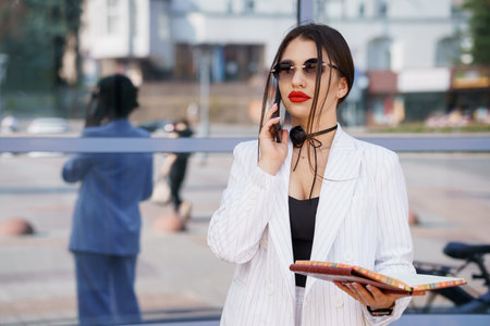 Chic Businesswoman in Stylish Striped Suit Engaged in Phone Call with Stylish Sunglasses and Notebook.の写真素材