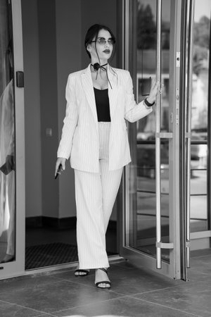 Stylish Businesswoman Exiting Elegant Glass Doors in Striped Suit and Sunglasses.の写真素材