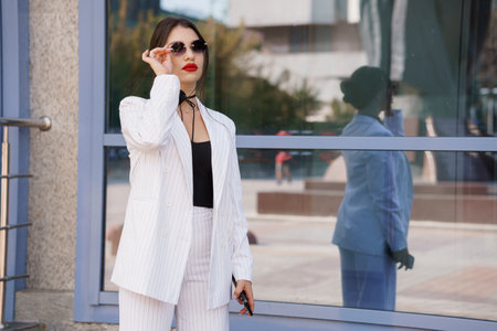 Stylish Woman in Striped Suit and Sunglasses Posing Confidently by Reflective Window.の写真素材