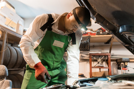 Mechanic in Green Overalls Working on Car Engine with Headlamp.の写真素材