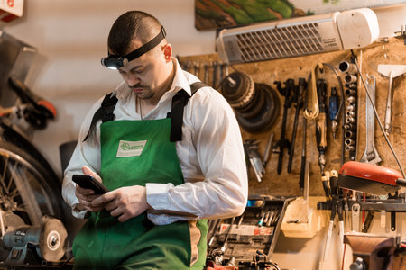 A Mechanic in a Green Apron Checking His Phone in a Workshop.