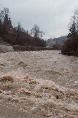 Turbulent River Flowing Under Overcast Skies Amidst Bare Trees and Hills.