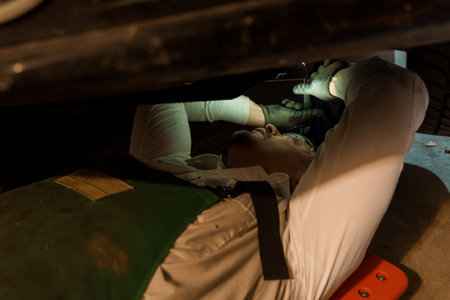 Professional Mechanic in Green Apron Inspecting Vehicle Underside with Flashlight in Repair Workshop.の写真素材