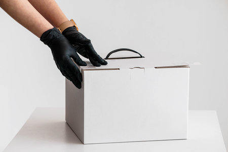 Hands in Black Gloves Carefully Closing White Cardboard Box on White Table.