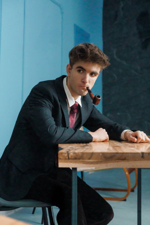 Young man in suit leaning on table with pipe in mouth, looking directly at viewer with serious expression.の写真素材