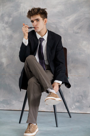 Young man in formal attire, wearing a blazer, tie, and trousers, sitting on a chair with legs crossed against a gray textured wall.の写真素材