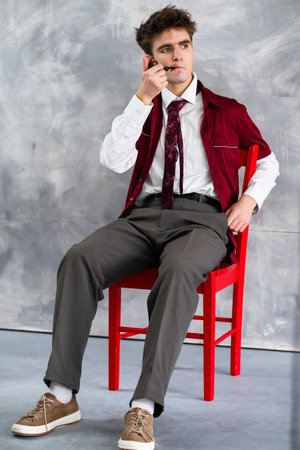 Young man sitting on a red chair while looking thoughtfully with stylish attire including burgundy...