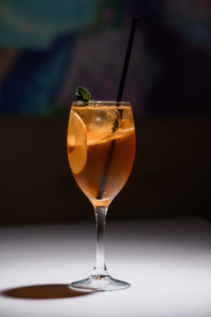 Refreshing cocktail in a wine glass with orange slice, ice cubes, mint garnish, and black straw on white surface with dark blurred background.の写真素材