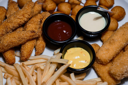 Appetizer platter with fried chicken strips, French fries, cheese curds and three dipping sauces on a white plate.の写真素材
