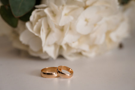 Wedding rings resting on a neutral surface surrounded by a lush bouquet of white hydrangeas and...