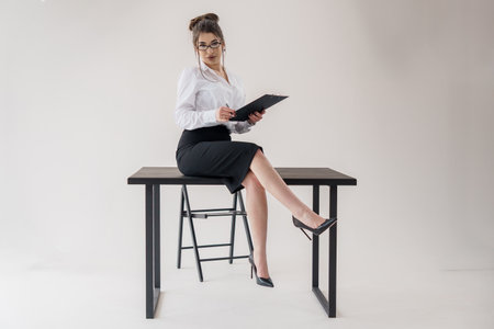 Confident businesswoman with clipboard seated on desk against neutral background.の写真素材