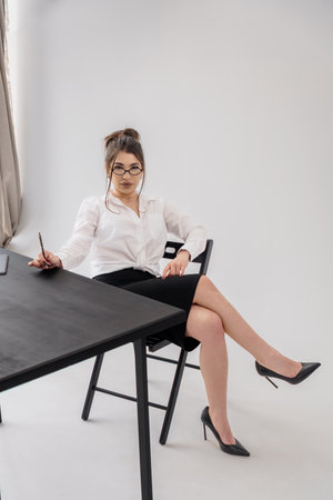 Focused businesswoman sits at black desk, holding pen with crossed legs.の写真素材