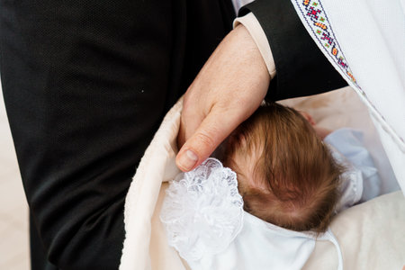 A tender moment of a baptism ceremony showcasing a man holding a baby wrapped in a white cloth with lace details, a priest's hand gently resting on the baby's head.の写真素材