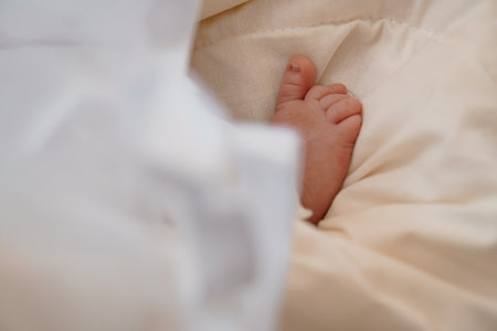 A close-up view of a newborn infant's tiny foot gently peeking out from under a soft, cream-colored blanket, symbolizing new life and tenderness with a parent's hand nearby.の写真素材
