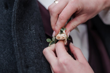 Hands carefully pin a soft peach-colored rose onto the charcoal gray fabric of a suit, symbolizing...