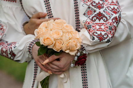 Close-up shows a couple holding a bouquet of peach roses during a traditional Ukrainian wedding, wearing intricately embroidered rusnyk shirts symbolizing loveの写真素材