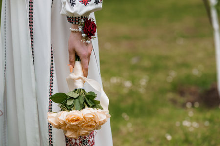 A close-up image shows a Ukrainian bride in a white vyshyvanka dress holding a delicate peach rose bouquet with a single red rose accent, representing love and traditionの写真素材