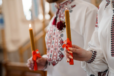 Two people hold beeswax candles decorated with red ribbons during an Orthodox wedding ritual,...