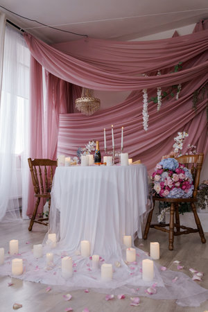 A beautifully arranged wedding table is set against a soft pink backdrop, adorned with elegant white linens, candles, floral arrangements, and a sparkling chandelier creating a dreamy atmosphereの写真素材
