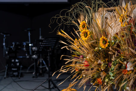 A vibrant floral arrangement of sunflowers, wheat, and dried grasses stands beside a drum set on a dimly lit stage, evoking a warm autumn harvest theme ideal for music or event backdropsの写真素材