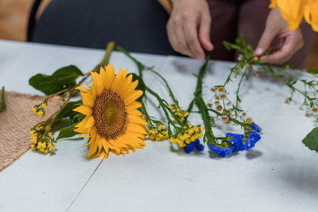A close-up view showcases hands arranging a vibrant floral display with sunflowers, blue statice, and yellow blossoms on a white wooden tableの写真素材
