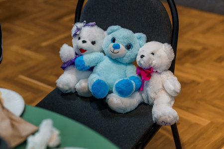 Three cute plush teddy bears one blue and two white are seated together on a black chair against a warm wooden floor, creating a heartwarming and cozy scene