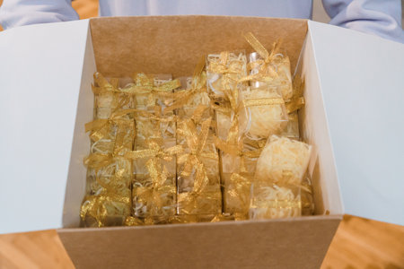 A cardboard box overflows with numerous small, individually wrapped favors tied with golden ribbons,...