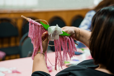 A person's hands are shown crafting a pink tassel decoration with a white flower at a table,...