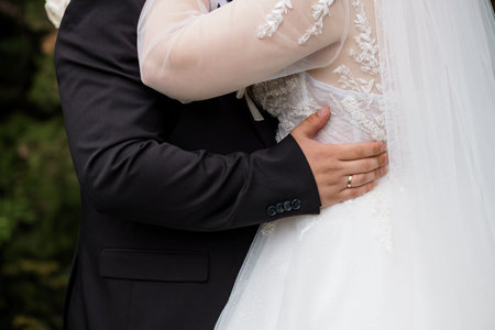 A tender wedding scene showing a groom closely embracing his bride, dressed in a beautiful lace wedding gown, wearing wedding rings, outdoors, joyful moodの写真素材