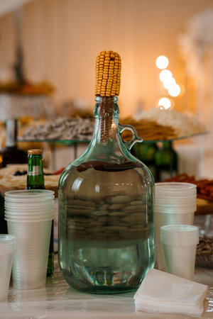An atmospheric image showing a green glass jug adorned with corn atop a table alongside white cups...