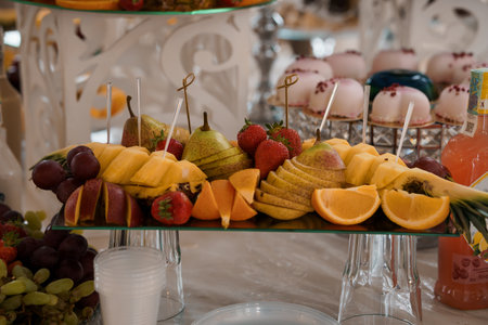 Luxurious fruit arrangement with oranges, pears, strawberries and grapes served at a wedding celebration. tiered stand, pink decorations, and glassware complement a festive moodの写真素材