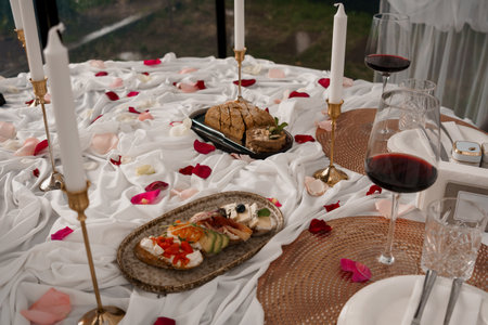 Intimate indoor picnic setting featuring wine glasses, cheese platters, bread, candles, and...